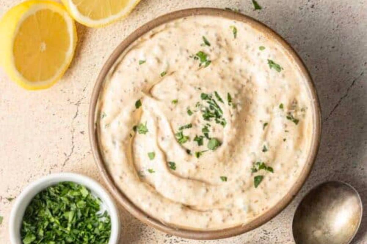 A bowl of creamy dip garnished with chopped herbs, perfect for pairing with next-level steak sauce or to sear it into your favorite dishes, sits beside lemon slices and a small bowl of more chopped herbs on a light surface.