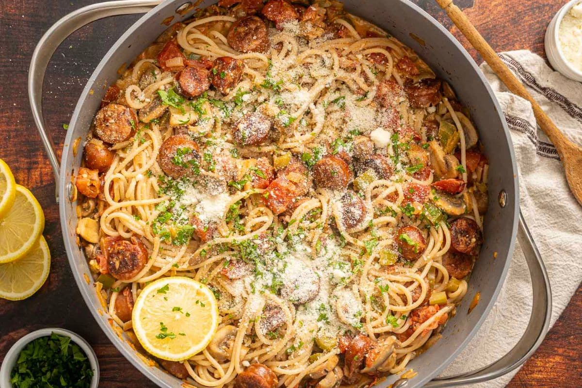 Large pan of spaghetti with sliced sausage, vegetables, tomato sauce, topped with grated cheese and fresh herbs-perfect for busy school nights. Garnished with lemon slices, this dish pairs well with any pork chop recipe. Wooden spoon and napkin nearby.