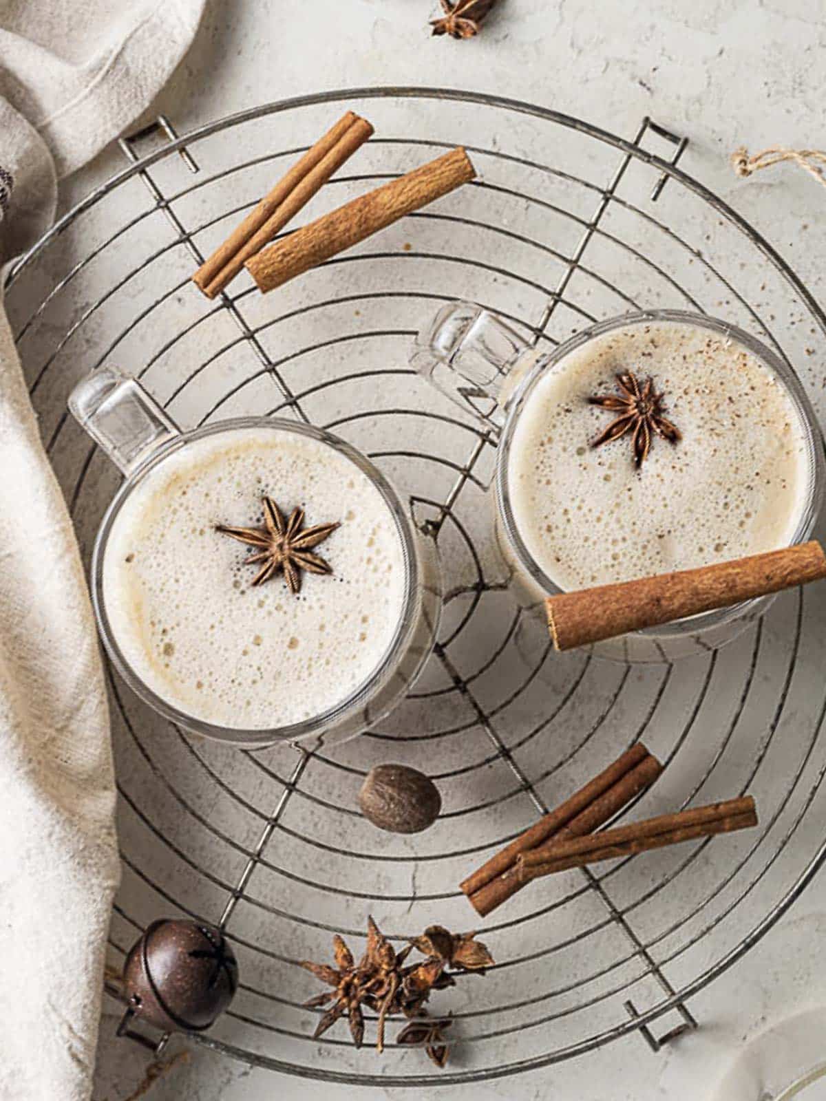 Two glass mugs of frothy chai latte topped with star anise, placed on a round cooling rack with cinnamon sticks and spices scattered around.