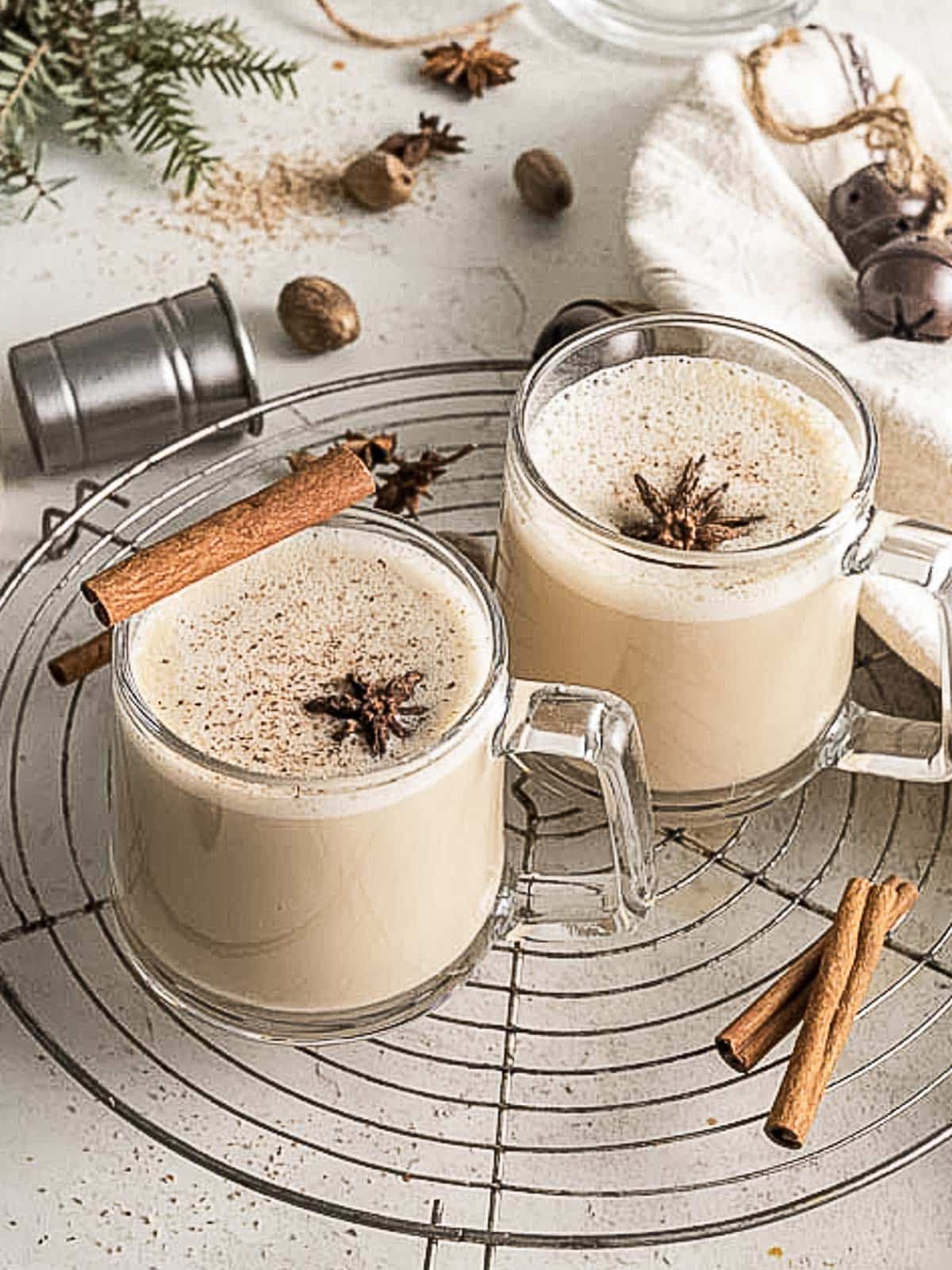 Two glass mugs of creamy eggnog topped with star anise and cinnamon sticks sit on a wire cooling rack, surrounded by spices and festive decor.