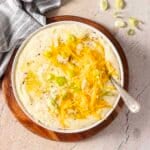 Bowl of mashed potatoes topped with shredded cheddar cheese, sliced green onions, and black pepper, with a spoon resting inside.