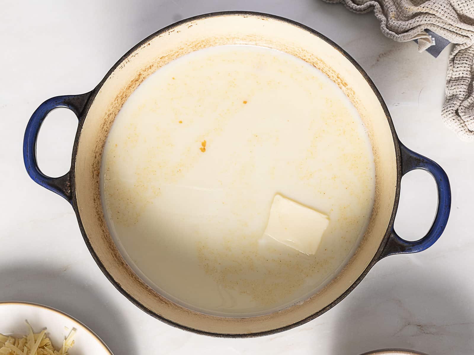 A blue-handled pot containing milk and a slab of butter, partially melted, sits on a light countertop with a towel nearby.