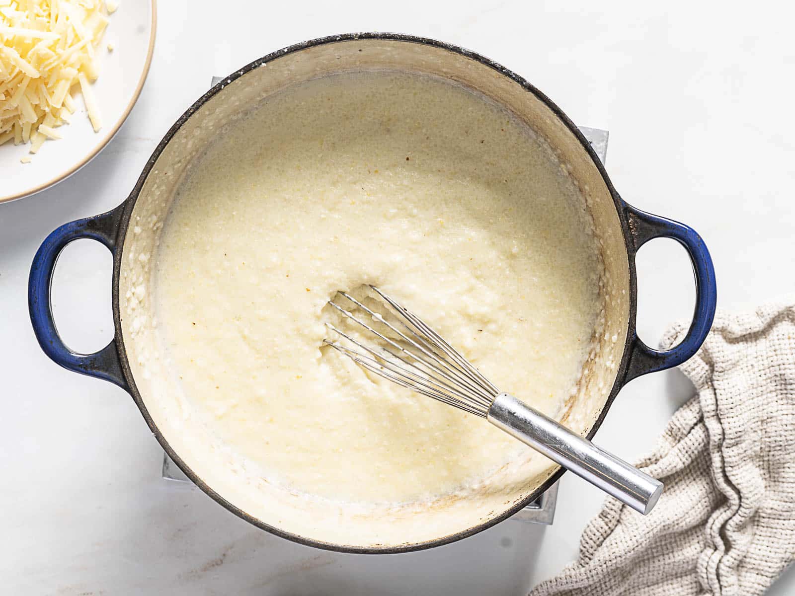 A pot of creamy mixture being whisked, with shredded cheese on a plate nearby and a textured cloth on the side.