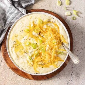 Bowl of mashed potatoes topped with shredded cheddar cheese, sliced green onions, and black pepper, with a spoon resting inside.