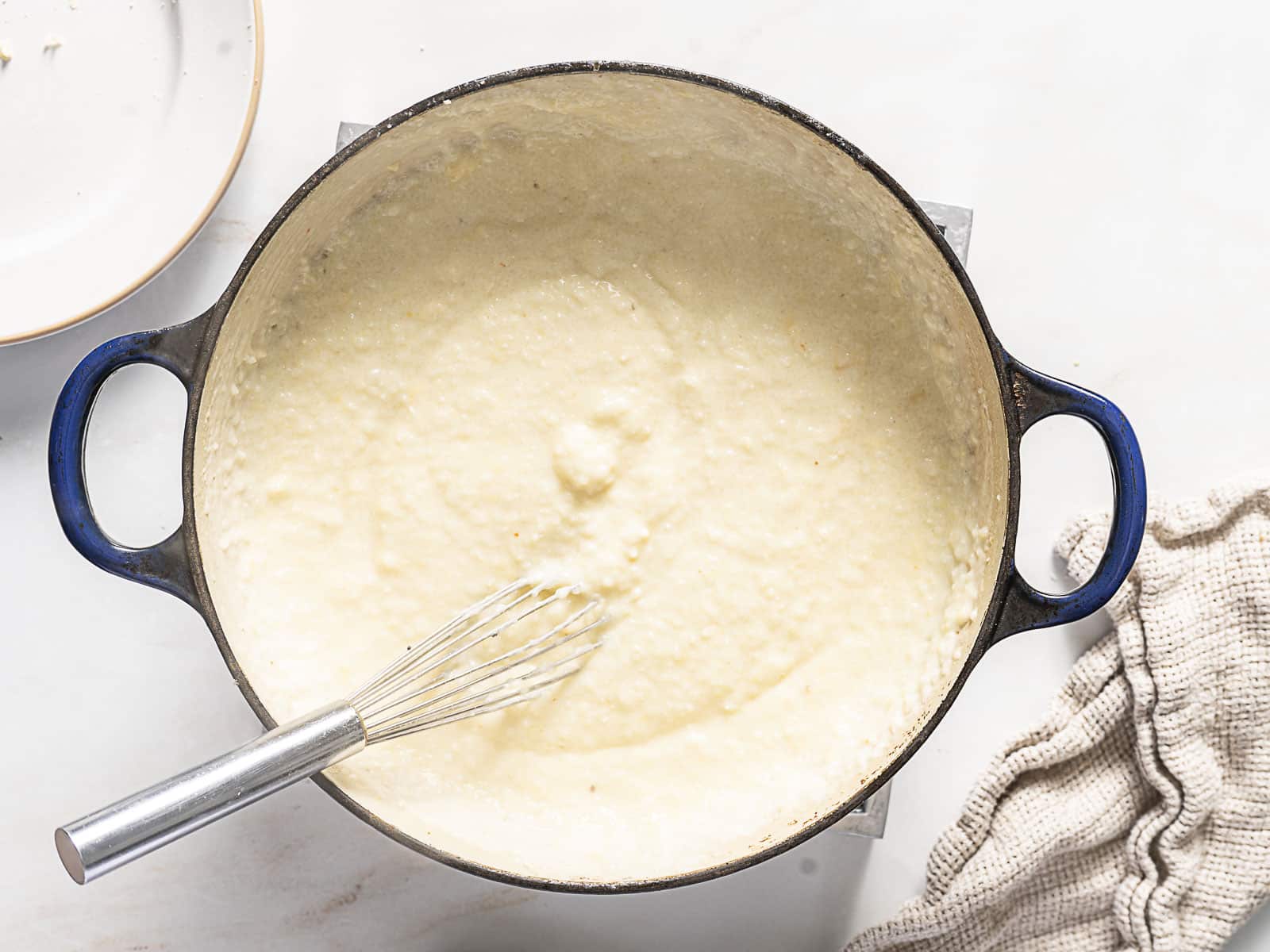 A pot of creamy grits is being stirred with a metal whisk, with a beige towel and plate nearby on a white surface.