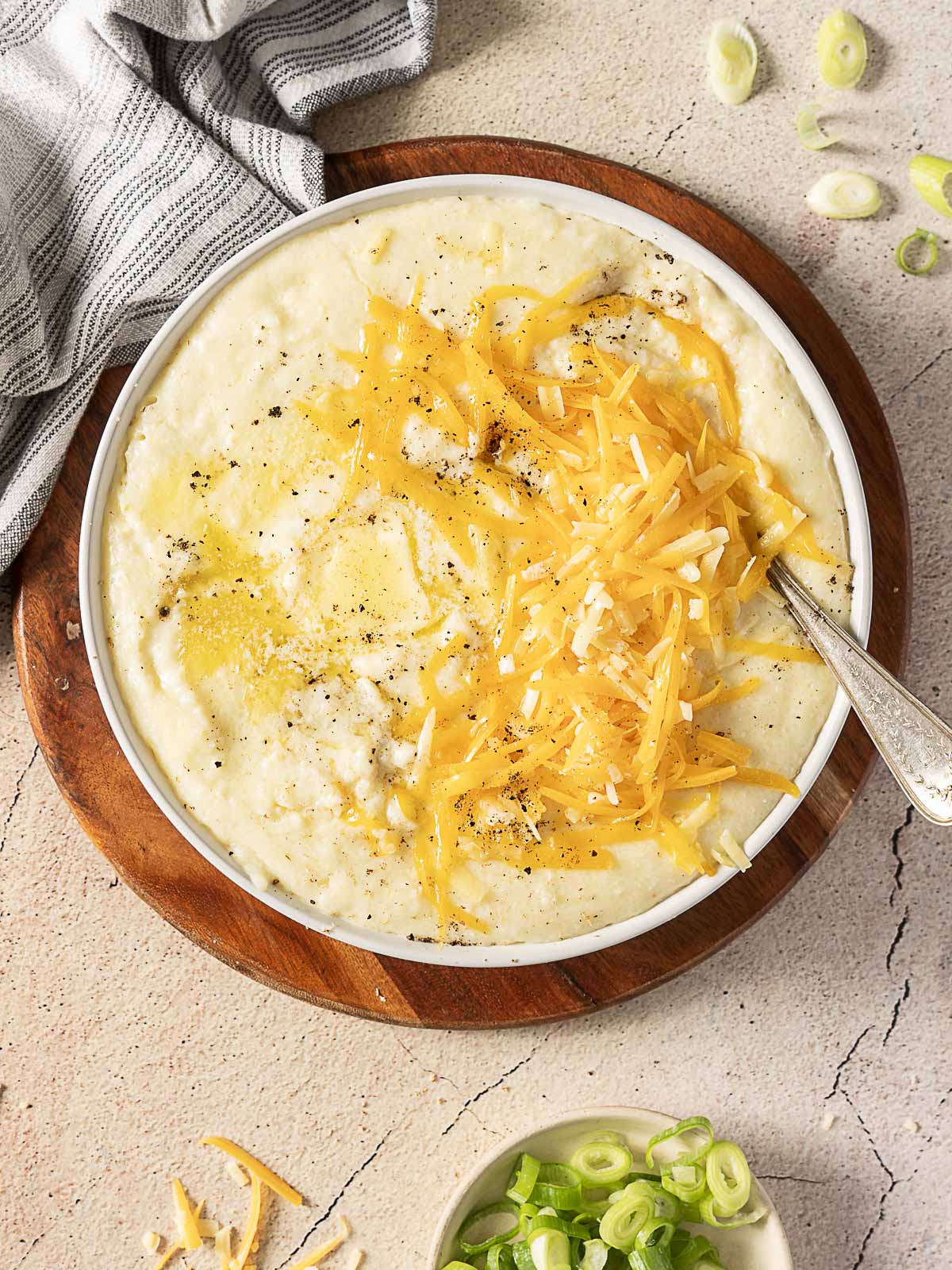 A bowl of creamy mashed potatoes is topped with shredded cheddar cheese and black pepper, with a spoon resting inside. Sliced green onions are in a small dish nearby.