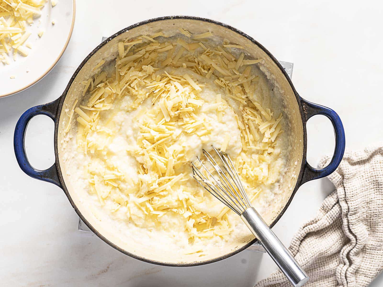 A pot of cheese sauce with shredded cheese and a whisk inside, sitting on a countertop next to a beige cloth and a plate of extra cheese.