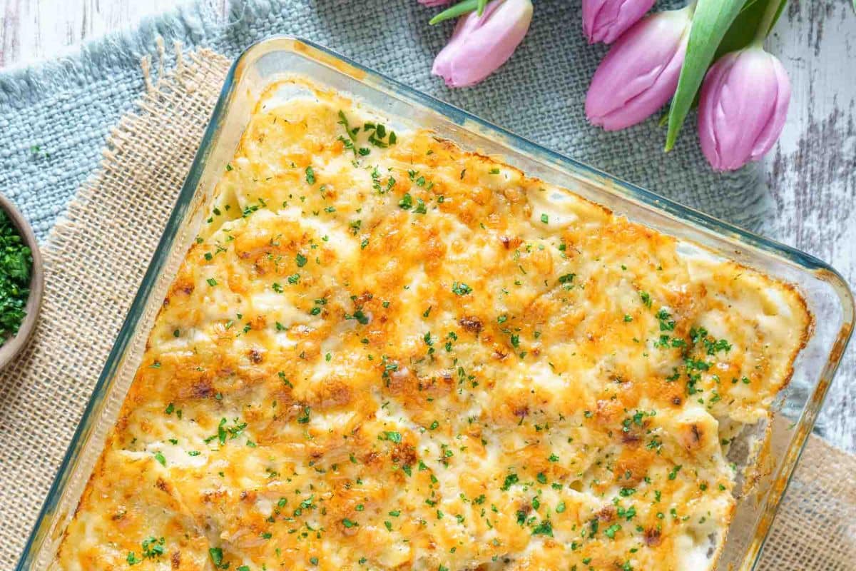 A glass baking dish filled with a cheesy baked casserole, topped with chopped herbs, sits on a textured cloth next to pink tulips-perfect for show-stealing side dishes at your next gathering.