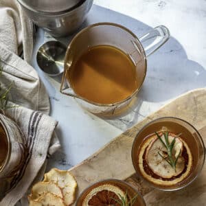 A glass pitcher of amber liquid sits on a marble surface next to two glasses garnished with dried citrus slices and herbs, a striped towel, and a metal measuring cup.