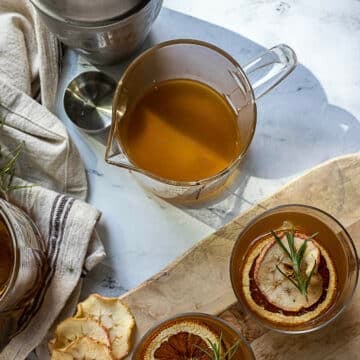 A glass pitcher of amber liquid sits on a marble surface next to two glasses garnished with dried citrus slices and herbs, a striped towel, and a metal measuring cup.