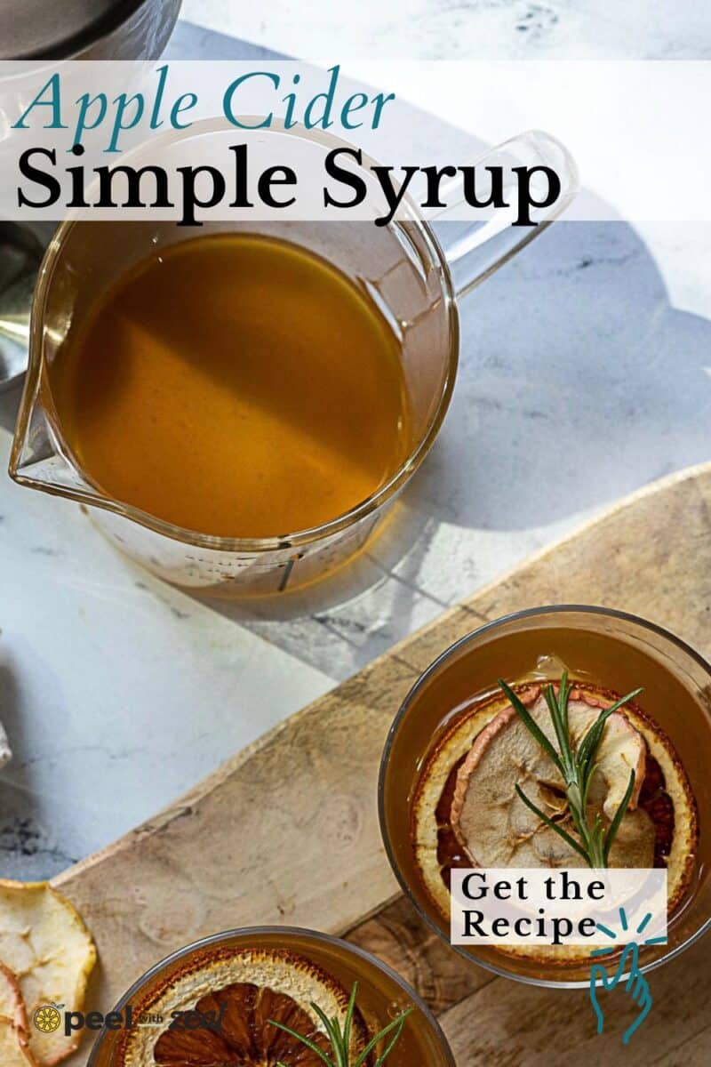 A glass measuring cup with cider syrup and two glasses of a drink garnished with dried apple slices and rosemary on a wooden board.