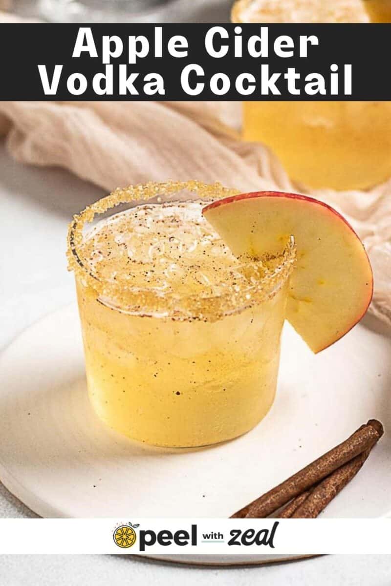 A glass of Apple Cider Moscow Mule with a sugar rim, garnished with an apple slice, sits on a white plate next to two cinnamon sticks.