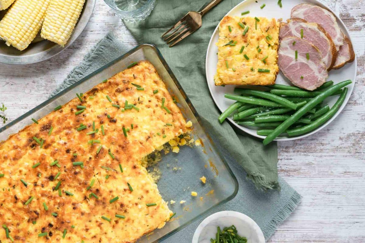 A glass baking dish of cornbread casserole with a serving removed; on the side, a plate with green beans, sliced ham, and a piece of casserole. Corn on the cob is in the background-pure comfort food for those who love soul-warming recipes.