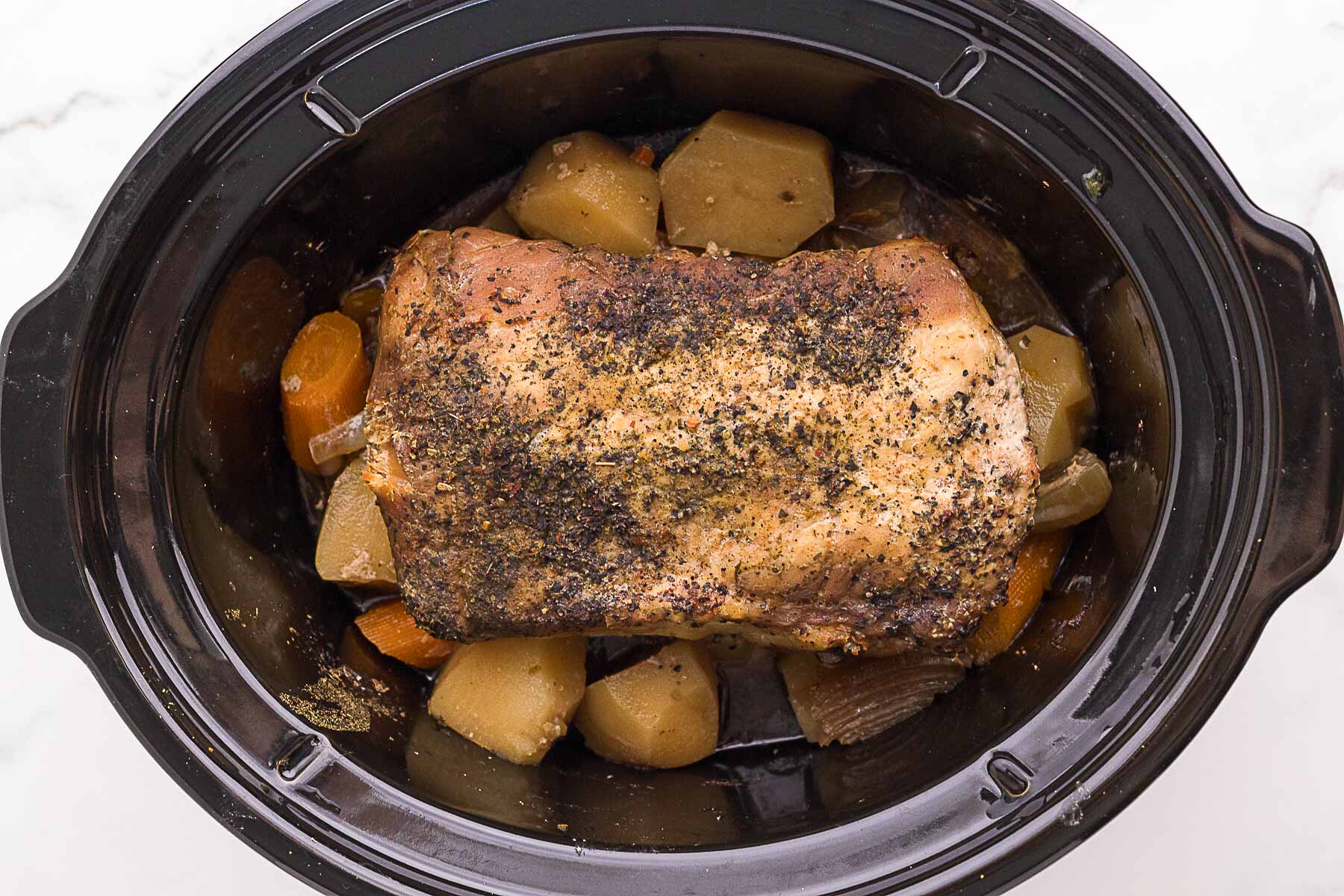 A seasoned pork roast sits on top of chopped potatoes, carrots, and onions inside a black slow cooker.