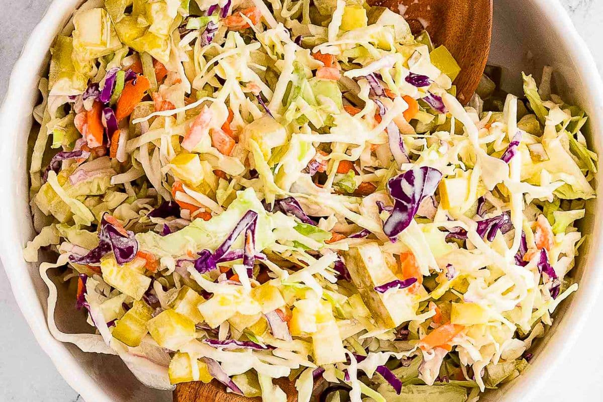 A close-up of a bowl of coleslaw with shredded cabbage, carrots, purple cabbage, pickles, and apple pieces in a creamy dressing-perfect for easy sides or as a fresh addition to your weeknight sides rotation.