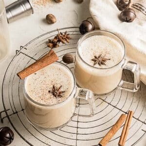 Two glass mugs of frothy chai latte topped with star anise and cinnamon sticks, placed on a cooling rack with spices scattered around.