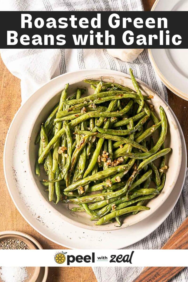 A bowl of garlic roasted green beans topped with chopped garlic sits on a wooden surface next to a small dish of pepper.