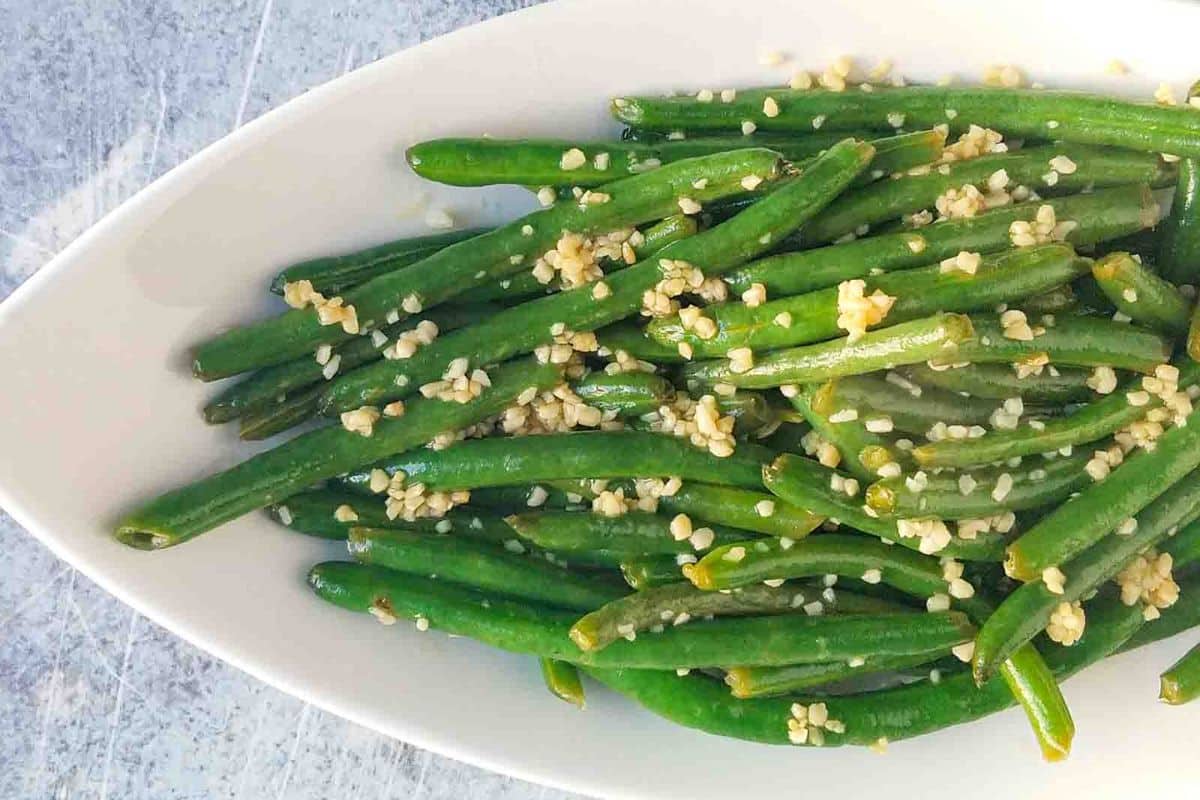 A white oval dish filled with cooked green beans topped with minced garlic-perfect for easy side dishes to elevate your weeknight sides.