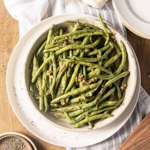 A bowl of cooked green beans garnished with minced garlic, placed on a striped cloth next to a stack of white plates and a small dish of pepper.