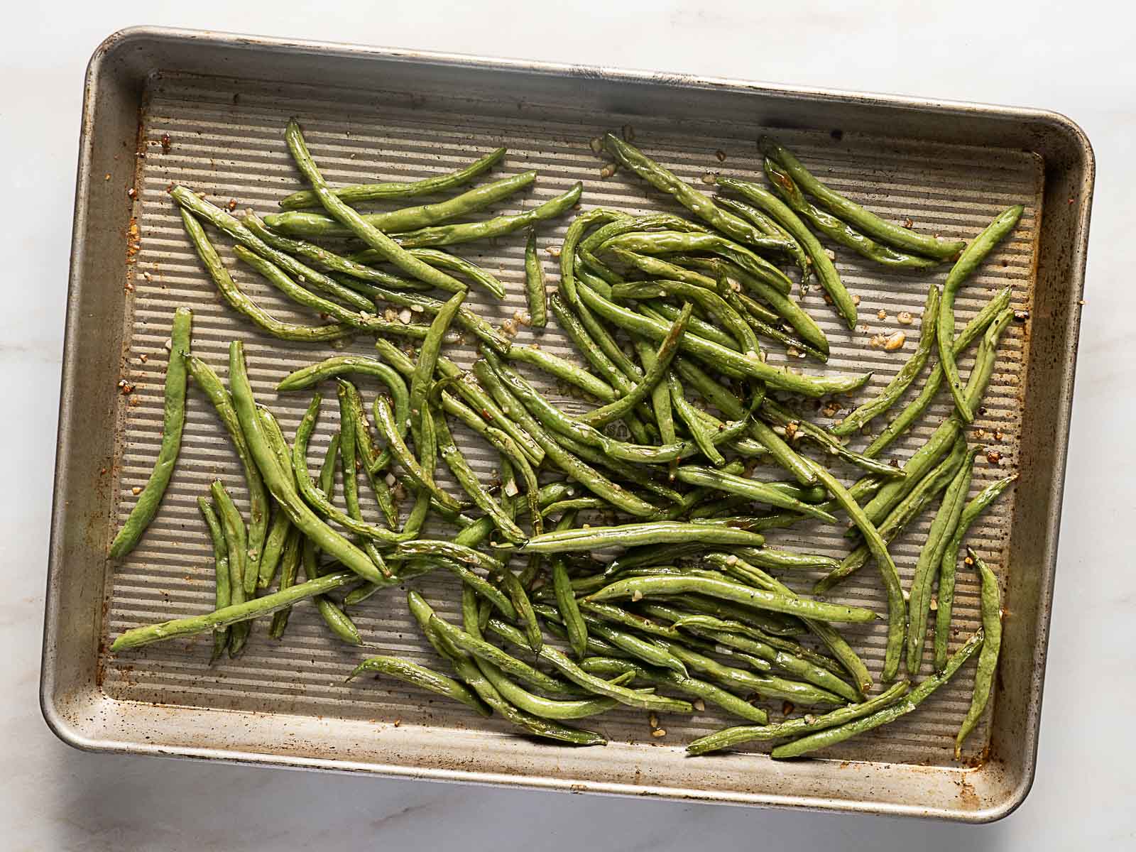 Roasted green beans spread out on a metal baking sheet, lightly browned and seasoned, on a white surface.