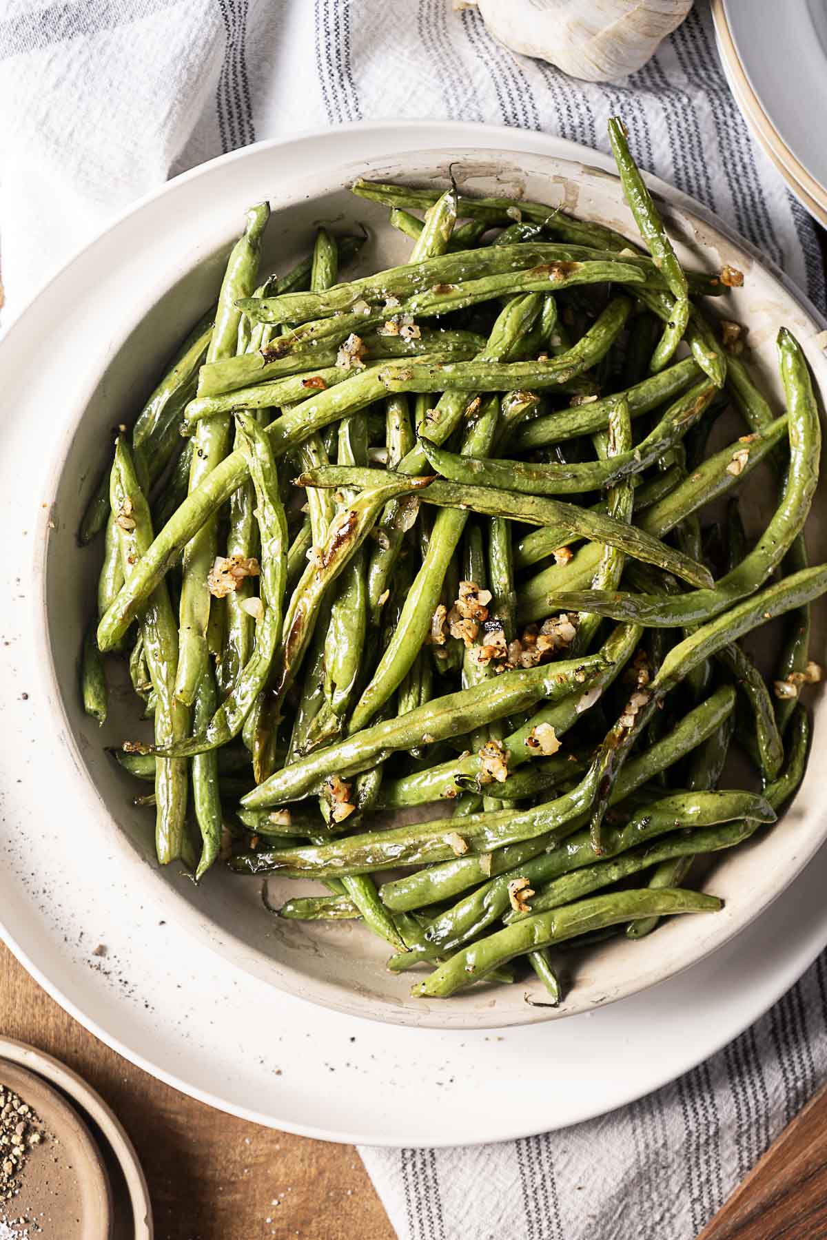 A bowl of cooked green beans garnished with minced garlic, served on a white plate with a striped cloth underneath.