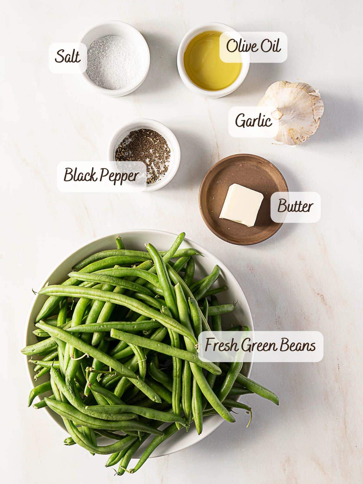 Ingredients for garlic roasted green beans arranged on a countertop: fresh green beans, salt, black pepper, olive oil, garlic, and a pat of butter.