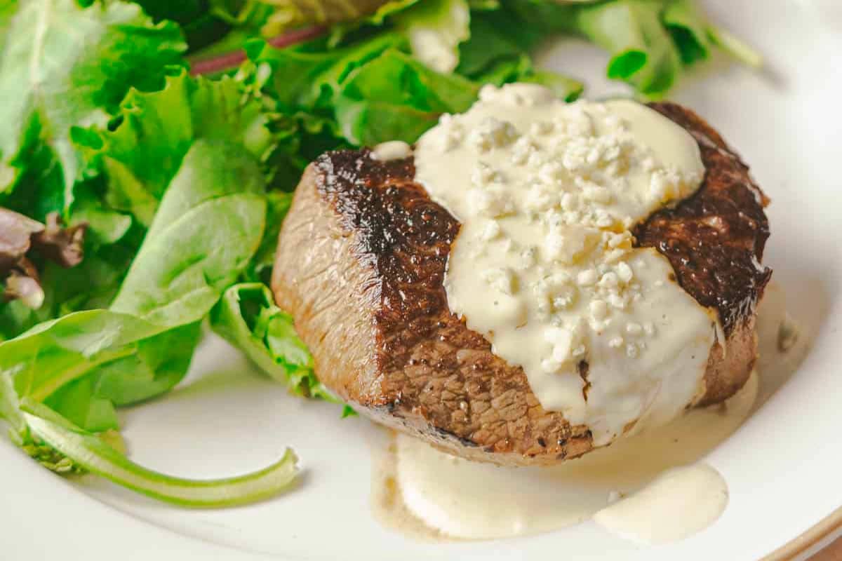 A grilled steak topped with creamy blue cheese sauce, inspired by classic steak sauce recipes, served alongside a fresh green salad on a white plate.