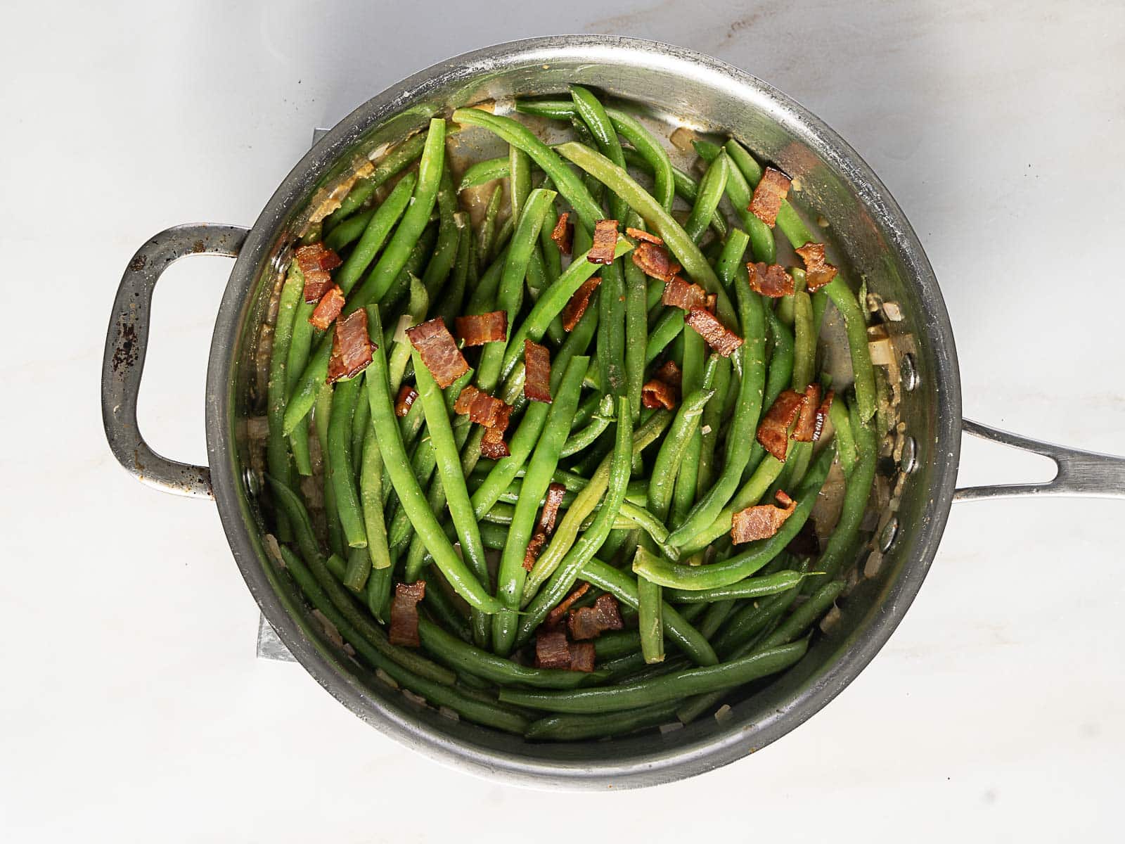 A stainless steel pan filled with cooked green beans topped with pieces of bacon, sitting on a light-colored surface.