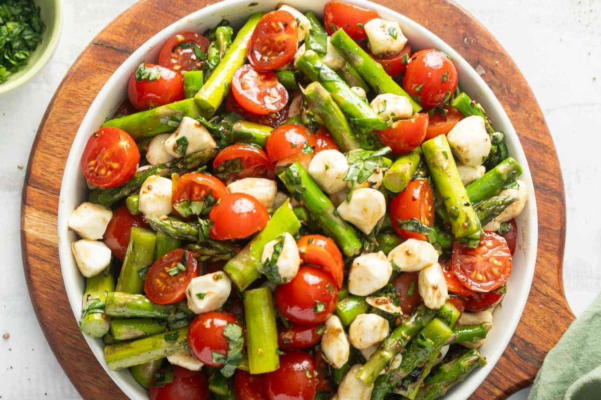 A bowl of salad with cherry tomatoes, asparagus, mozzarella balls, and herbs on a wooden board-one of the best side dishes that's sure to be a show-stealing side at any meal.