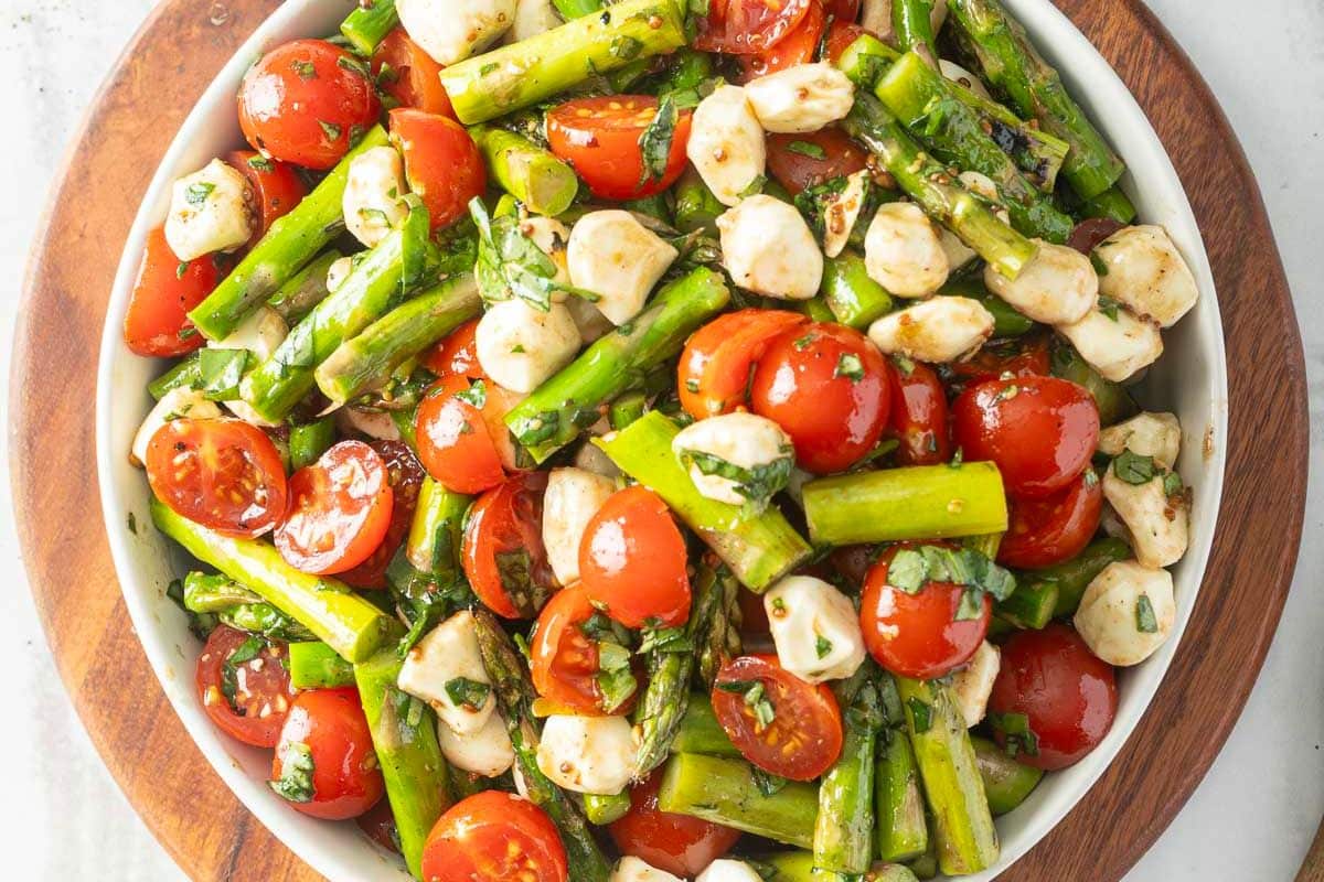 A bowl of salad with cherry tomatoes, asparagus, mozzarella balls, and chopped herbs on a wooden surface-perfect for easy side dishes or weeknight sides.