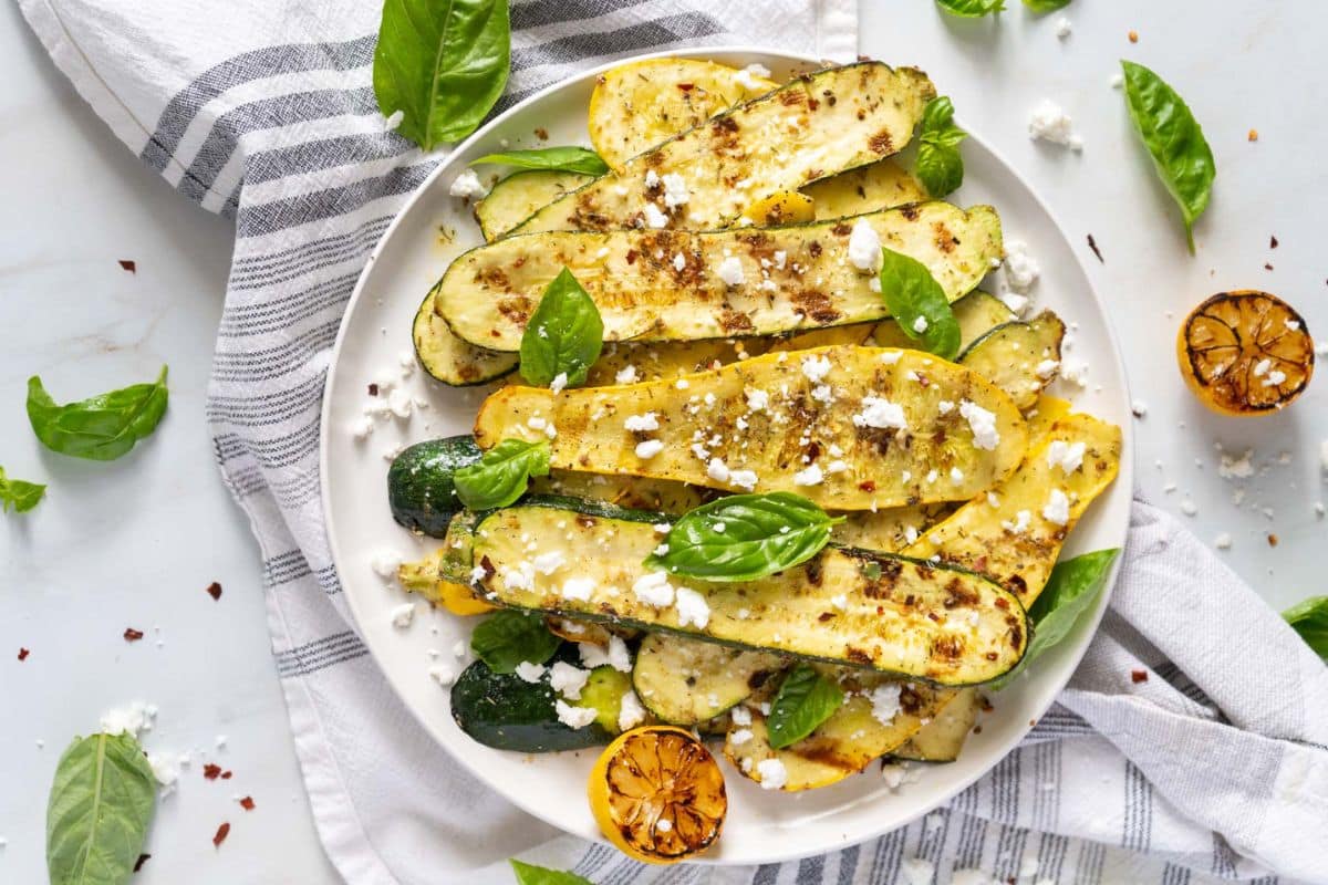 A plate of grilled zucchini and yellow squash topped with crumbled cheese and herbs, garnished with basil leaves and served with charred lemon halves-one of our favorite easy weeknight recipes.