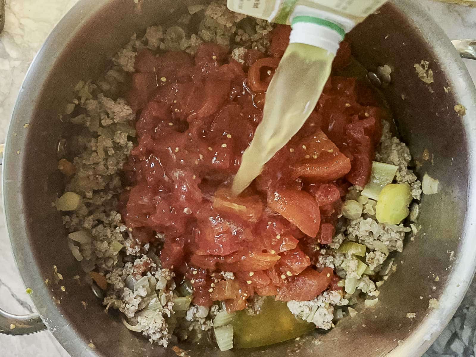 Ground meat, chopped vegetables, and canned tomatoes in a pot with broth being poured in, viewed from above on a kitchen countertop.