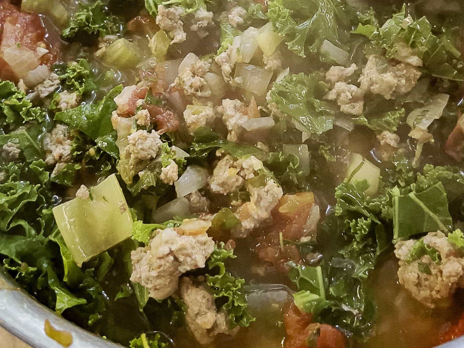 A close-up of a pot of soup containing ground meat, chopped kale, diced tomatoes, onions, and celery in broth.
