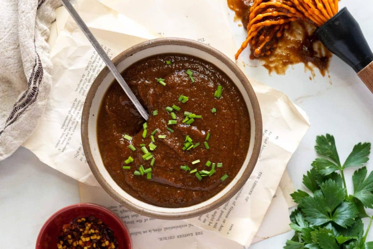 A bowl of rich, dark brown steak sauce garnished with chopped chives sits next to a spoon, herbs, red pepper flakes, and a sauce-covered whisk on a white surface. Perfect for elevating your next-level steak sauce recipes.
