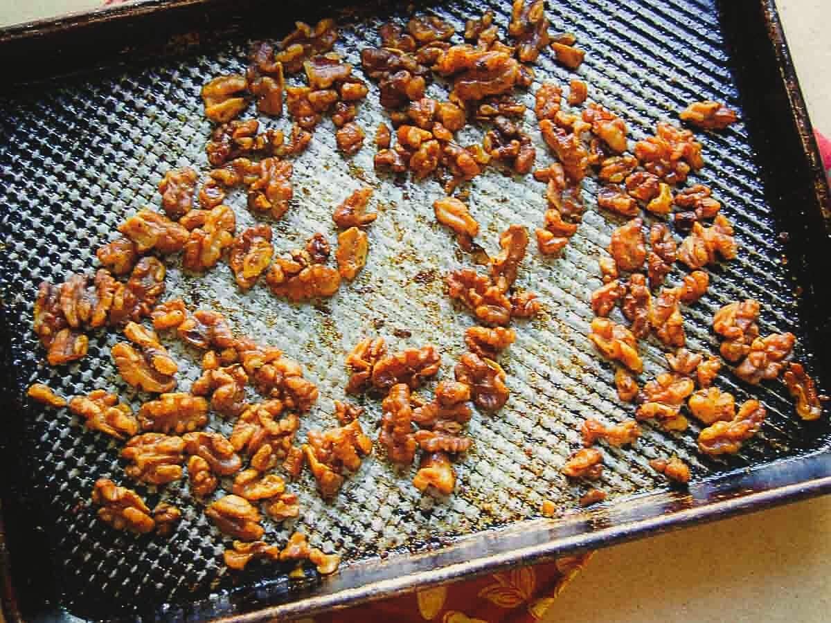 Walnut halves coated in a glaze are spread out on a textured baking sheet, some areas showing caramelization.