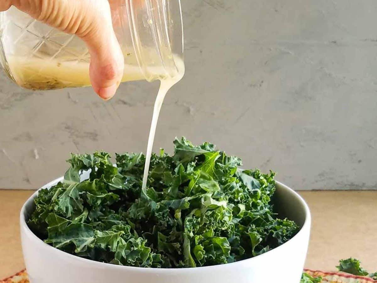 A hand pours creamy dressing from a jar over a bowl of fresh kale leaves.
