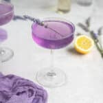 A purple cocktail in a coupe glass garnished with a sprig of lavender, with a halved lemon and additional lavender sprigs in the background.
