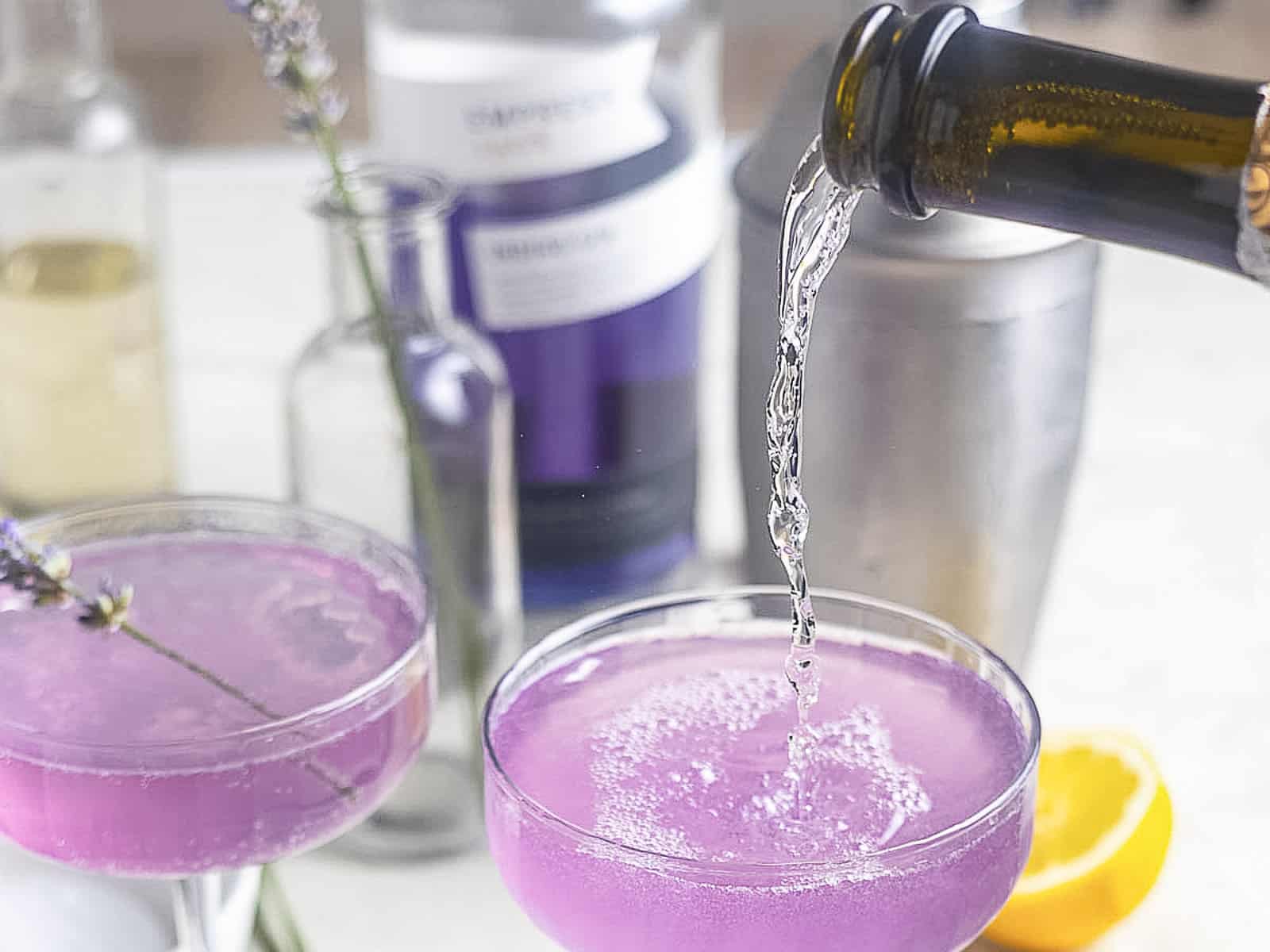 A clear liquid is being poured into a coupe glass with a purple cocktail. Bottles, a shaker, and a lemon half are in the background.
