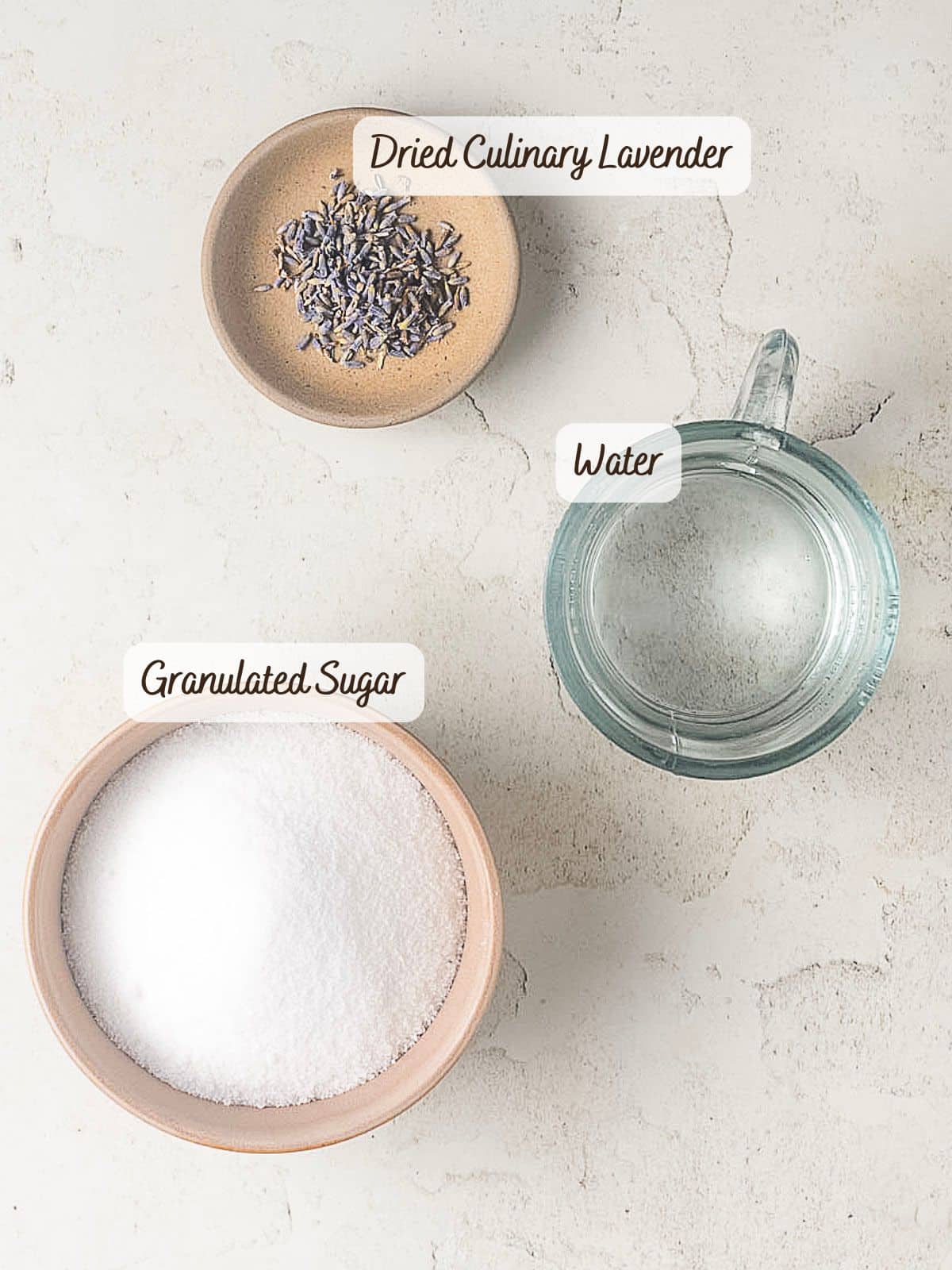 A bowl of dried culinary lavender, a glass measuring cup of water, and a bowl of granulated sugar are arranged on a light surface&mdash;perfect ingredients for making homemade lavender simple syrup.
