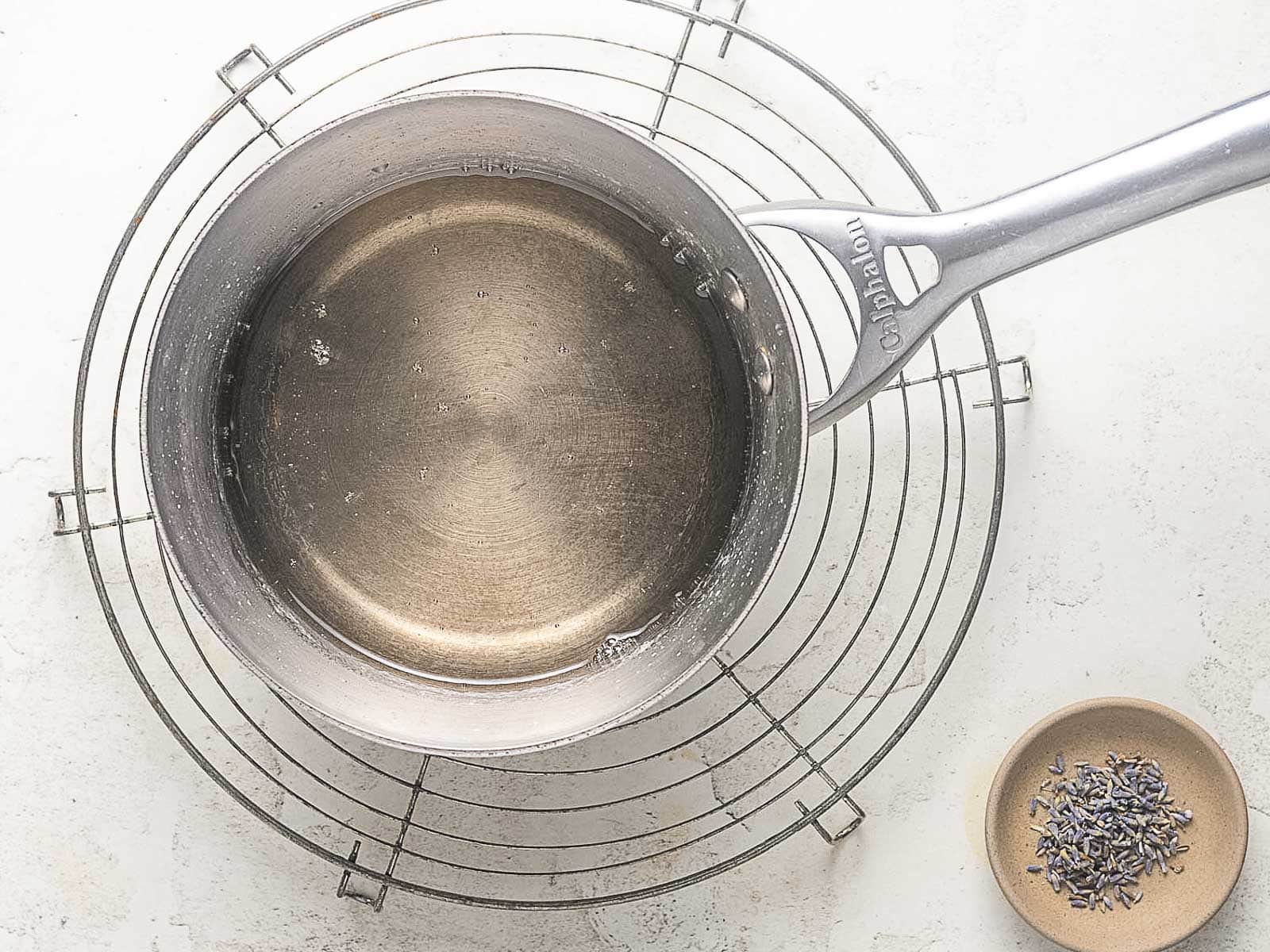 A metal saucepan with clear liquid sits on a round cooling rack next to a small dish of dried lavender buds on a white surface.