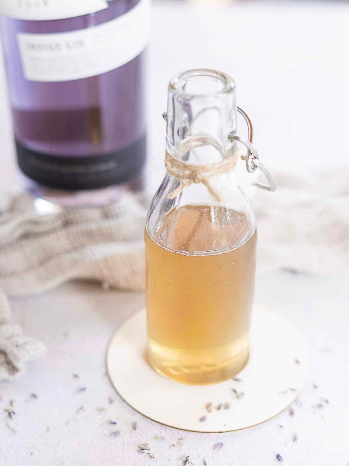 A small glass bottle filled with light brown lavender simple syrup sits on a round coaster, with a bottle and cloth in the blurred background. Lavender buds are scattered on the surface.