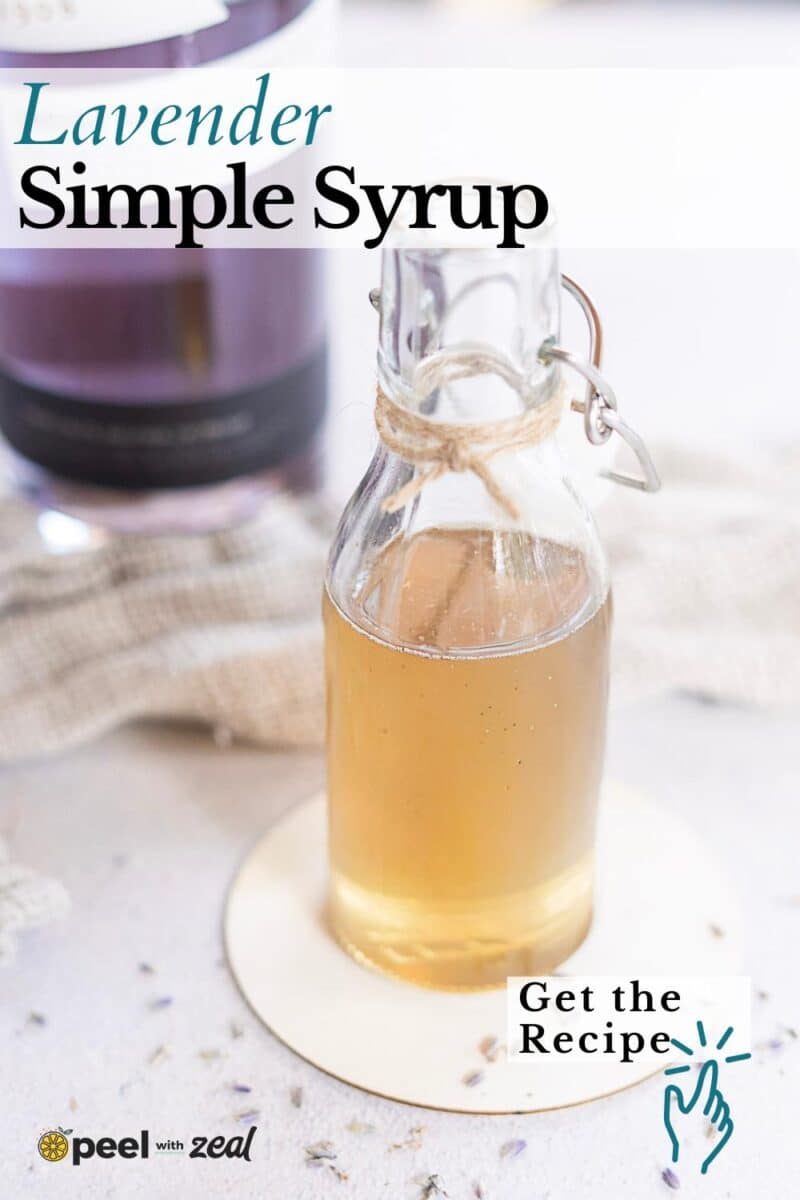 A small glass bottle of lavender simple syrup sits on a coaster, surrounded by scattered lavender buds, with a blurred bottle in the background.