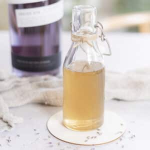 A small glass bottle filled with lavender simple syrup sits on a coaster, with a blurred purple-labeled bottle and a textured cloth in the background.
