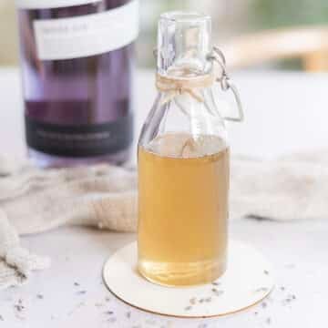 A small glass bottle filled with lavender simple syrup sits on a coaster, with a blurred purple-labeled bottle and a textured cloth in the background.
