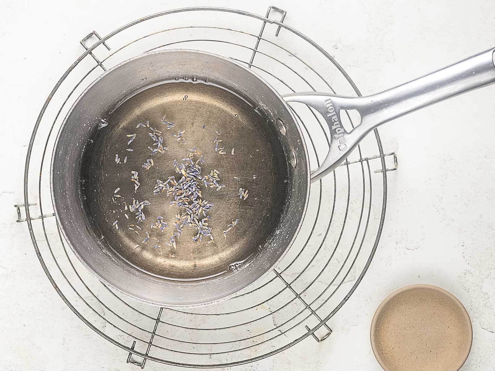 A metal saucepan with water and dried lavender sits on a wire rack next to a small empty ceramic bowl on a white surface.