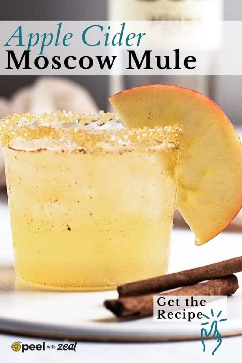 A glass of Apple Cider Moscow Mule garnished with a fresh apple slice and a sugared rim, with two cinnamon sticks in the foreground.