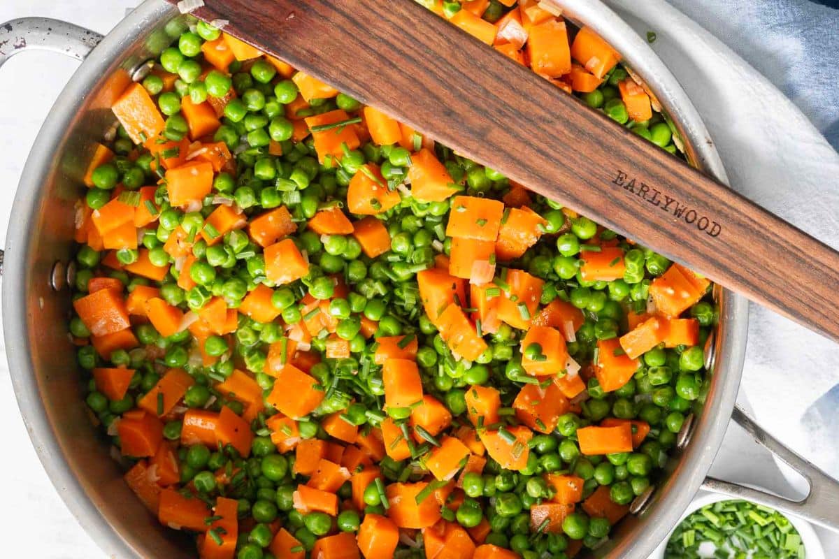 A pan filled with cooked diced carrots and green peas, mixed together, with a wooden spatula resting on top-one of those easy side dishes perfect for weeknight sides.