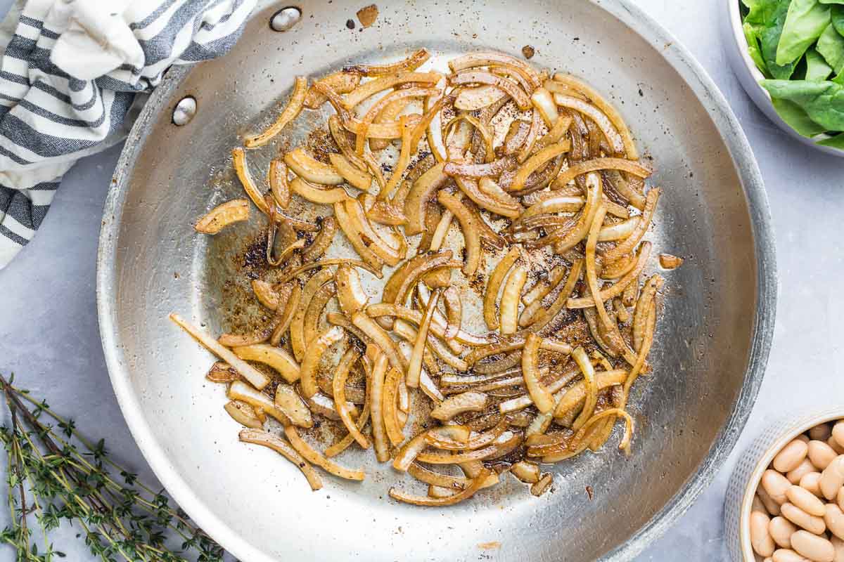 Saut&eacute;ed sliced onions browning in a stainless steel pan, with a napkin, fresh herbs, spinach, and white beans nearby-perfect for School Night Dinners or pairing with your favorite Easy Pork Recipes.