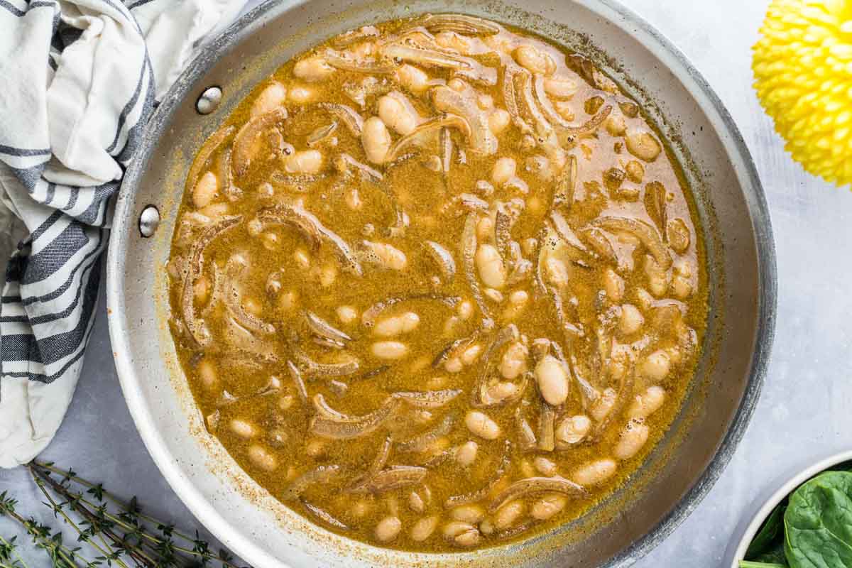 A pan filled with a brown broth containing white beans and shredded pieces of meat makes an ideal School Night Dinner, with a striped cloth and a yellow flower nearby.