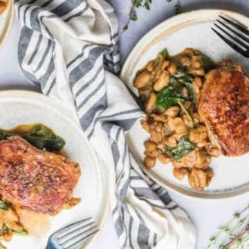 Two plates with skillet pork chops served over sautéed white beans and spinach, set beside a striped napkin and sprigs of thyme—an easy dinner that's both hearty and flavorful.